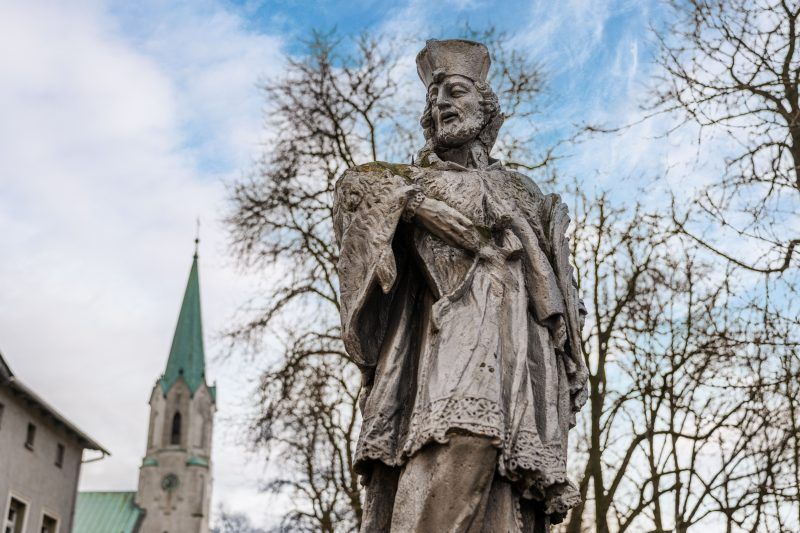 Na zdjęciu widać kamienną figurę św. Jana Nepomucena z krzyżem w dłoniach, stojącą na ozdobnym cokole.