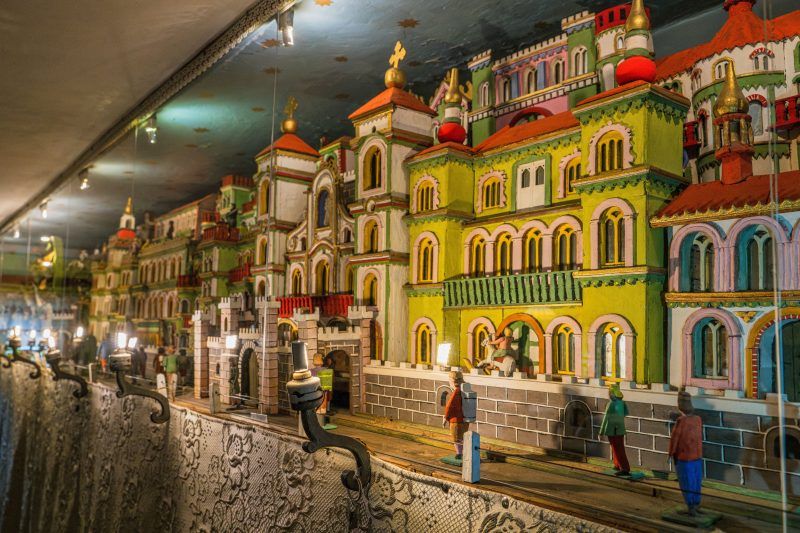 Na zdjęciu widać wnętrze drewnianego domku wypełnione ruchomą szopką – rzędy misternie wyrzeźbionych, kolorowych figurek poruszają się wśród miniaturowych domków, drzew i scen biblijnych, tworząc ciepłą, rękodzielniczą atmosferę.