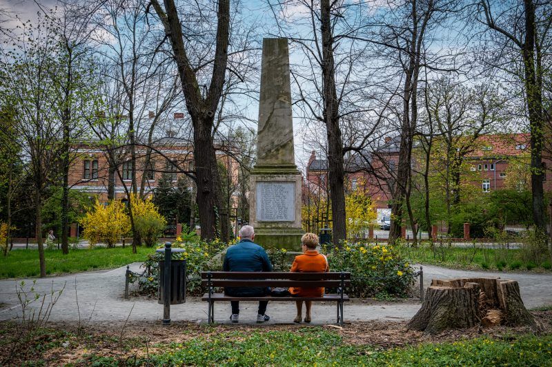 Kamienny obelisk stoi pośród zieleni parku, otoczony drzewami i spokojną alejką.