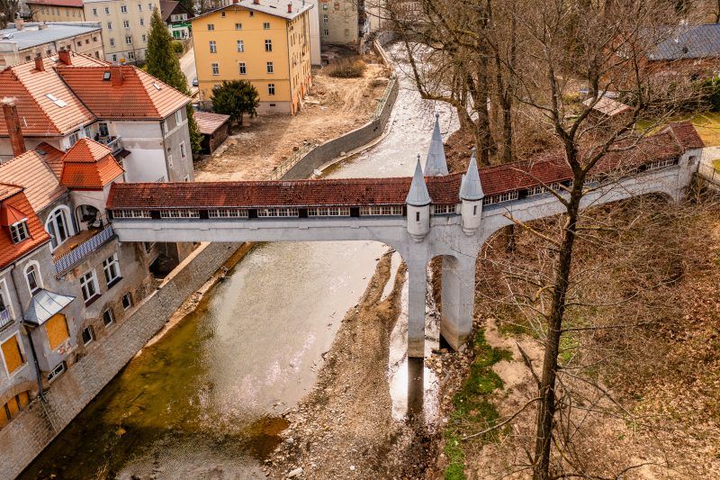 Kryty Most w Lądku-Zdroju to zabytkowa kamienna konstrukcja z łukowym przejściem, przykryta drewnianym dachem, otoczona bujną roślinnością i malowniczym krajobrazem.
