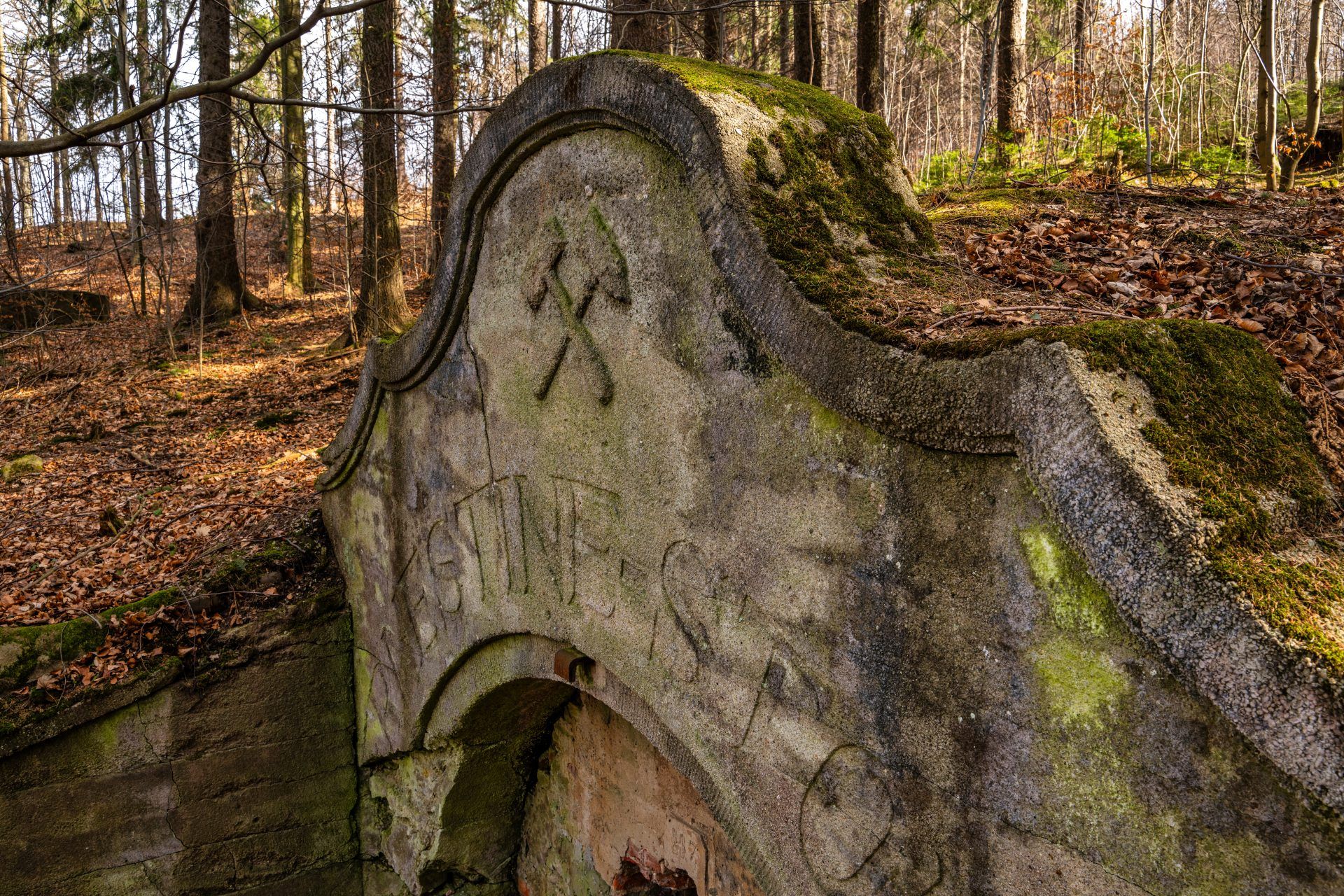 Na zdjęciu widać kamienny portal wejściowy sztolni Ernestine, otoczony przez zarośnięte wzgórze, z surowymi skałami i ścianą góry w tle.