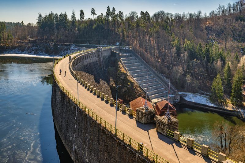 Na zdjęciu widać potężną kamienną zaporę w Pilchowicach, majestatycznie rozciągającą się wśród gór i odbijającą się w spokojnej tafli jeziora.