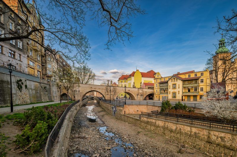 Na zdjęciu widać stary, kamienny most ozdobiony barokowymi rzeźbami świętych, rozciągający się łagodnym łukiem nad rzeką, z tle malowniczymi kamieniczkami kłodzkiej starówki.