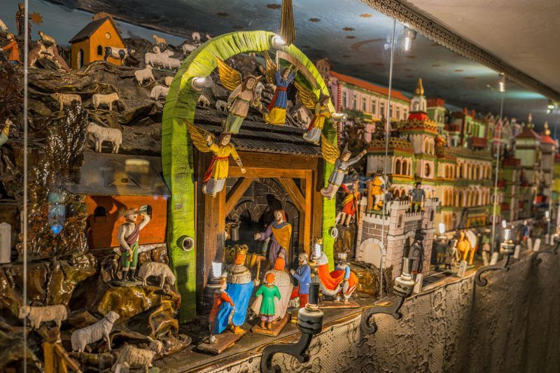 Na zdjęciu widać wnętrze drewnianego domku wypełnione ruchomą szopką – rzędy misternie wyrzeźbionych, kolorowych figurek poruszają się wśród miniaturowych domków, drzew i scen biblijnych, tworząc ciepłą, rękodzielniczą atmosferę.