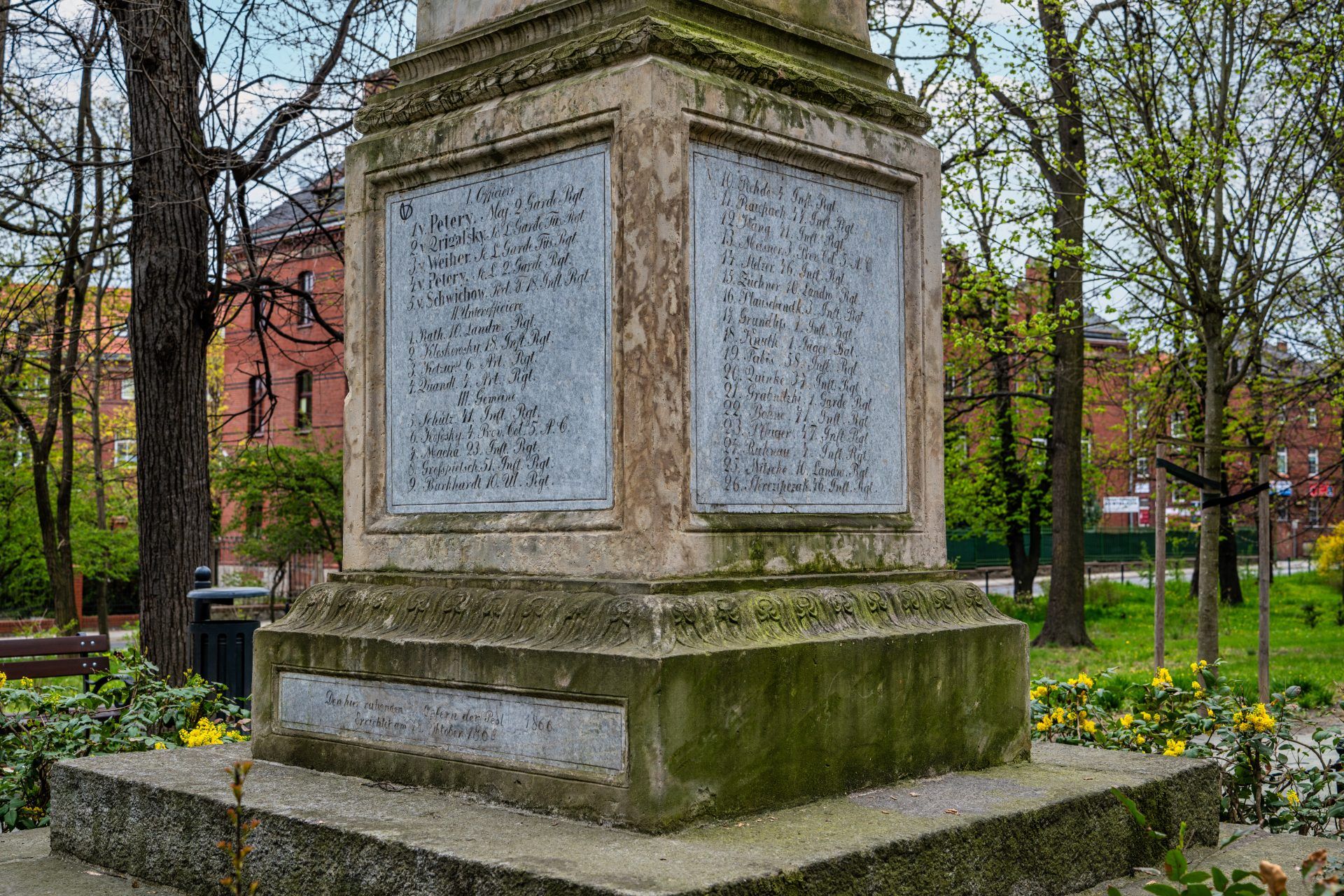 Kamienny obelisk stoi pośród zieleni parku, otoczony drzewami i spokojną alejką.