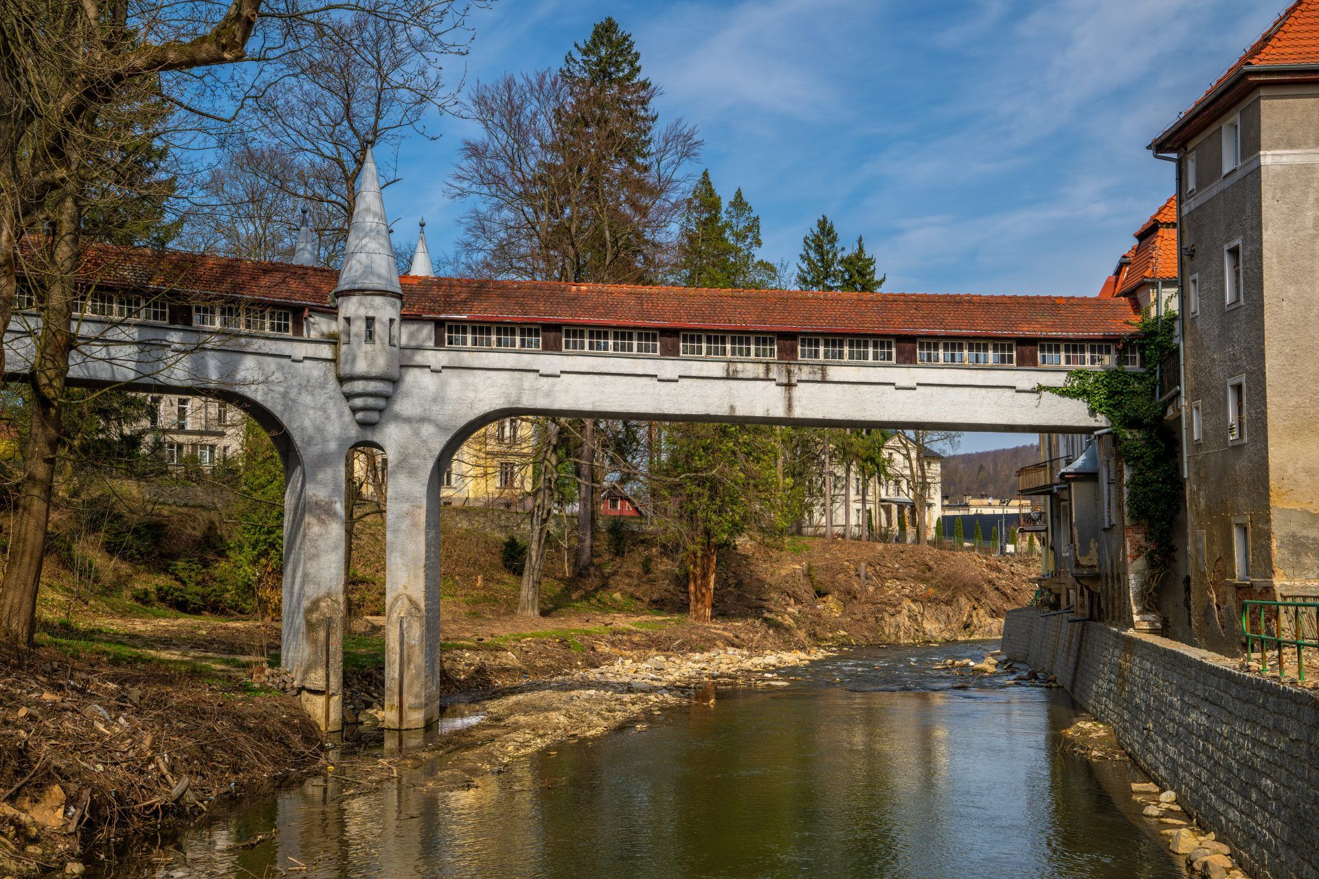 Kryty Most w Lądku-Zdroju to zabytkowa kamienna konstrukcja z łukowym przejściem, przykryta drewnianym dachem, otoczona bujną roślinnością i malowniczym krajobrazem.