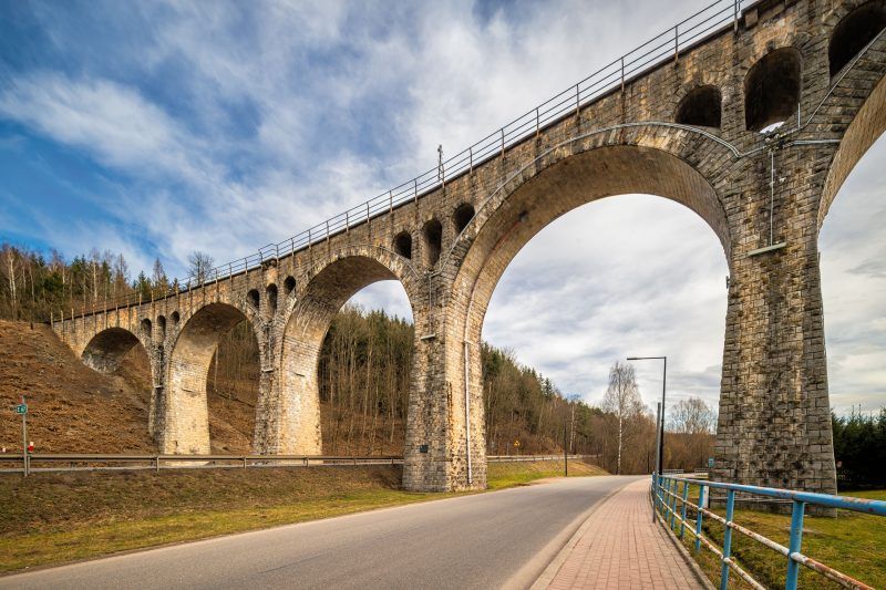 Na zdjęciu widać masywny kamienny wiadukt z łukami, wznoszący się nad doliną, otoczony bujną roślinnością i malowniczym krajobrazem.