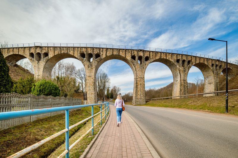 Na zdjęciu widać masywny kamienny wiadukt z łukami, wznoszący się nad doliną, otoczony bujną roślinnością i malowniczym krajobrazem.