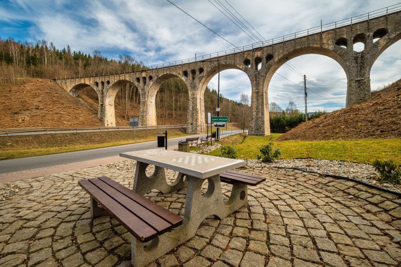 Na zdjęciu widać masywny kamienny wiadukt z łukami, wznoszący się nad doliną, otoczony bujną roślinnością i malowniczym krajobrazem.