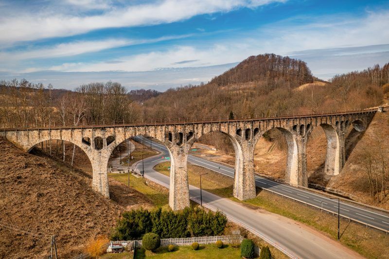 Na zdjęciu widać masywny kamienny wiadukt z łukami, wznoszący się nad doliną, otoczony bujną roślinnością i malowniczym krajobrazem.