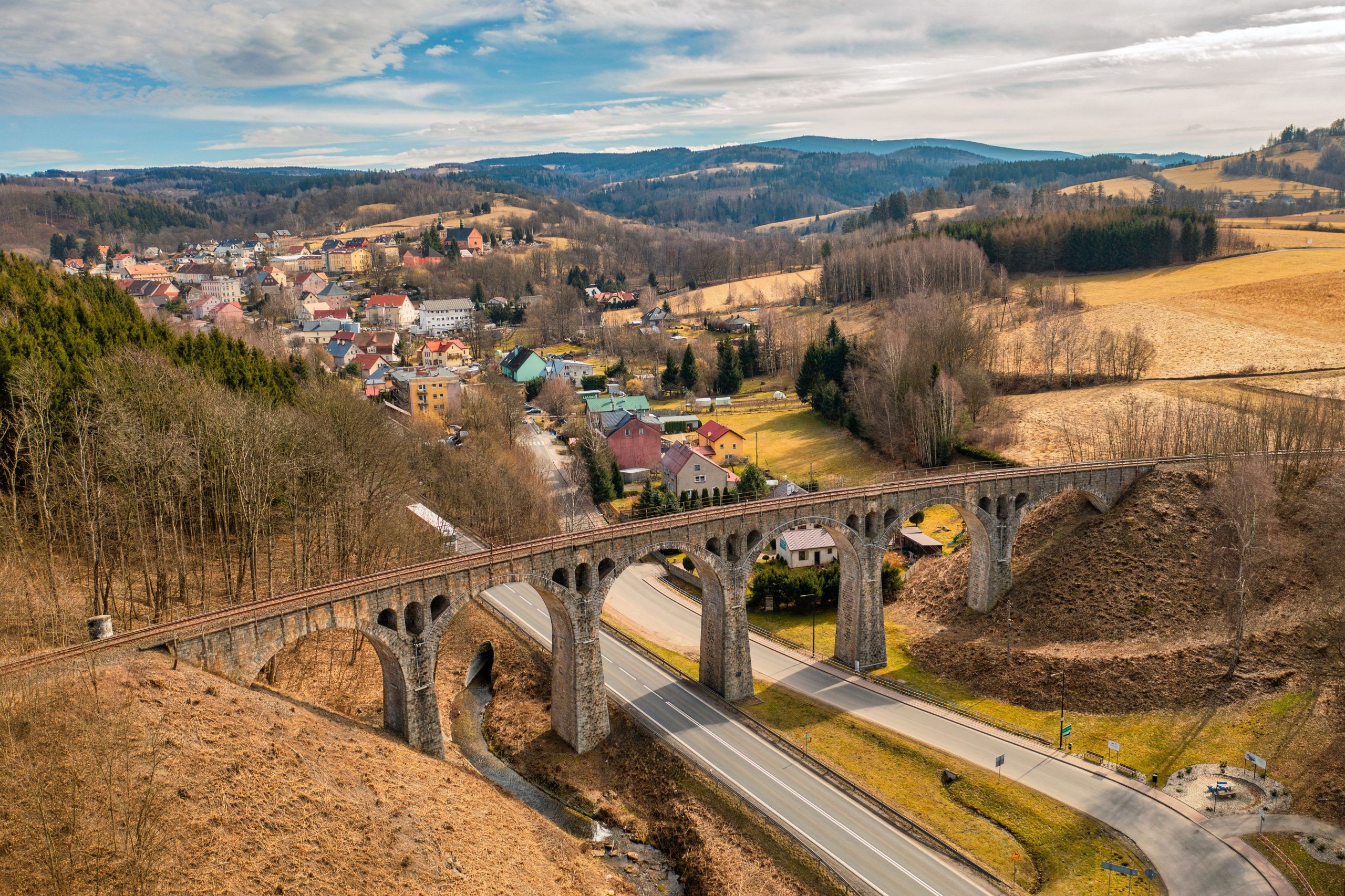 Na zdjęciu widać masywny kamienny wiadukt z łukami, wznoszący się nad doliną, otoczony bujną roślinnością i malowniczym krajobrazem.