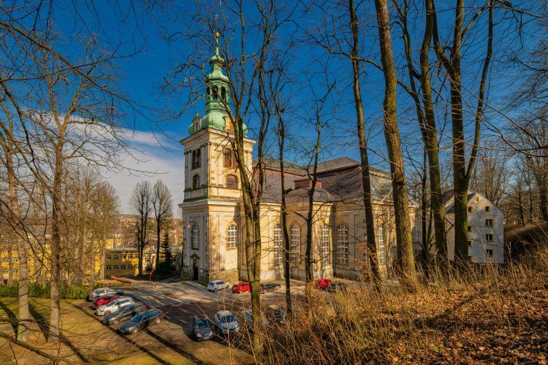 Na zdjęciu widać okazały Kościół Łaski w Kamiennej Górze – barokową budowlę o jasnej elewacji, wysokim dachu i smukłej wieży górującej nad otaczającym ją parkiem.