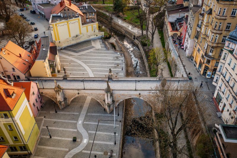 Na zdjęciu widać stary, kamienny most ozdobiony barokowymi rzeźbami świętych, rozciągający się łagodnym łukiem nad rzeką, z tle malowniczymi kamieniczkami kłodzkiej starówki.