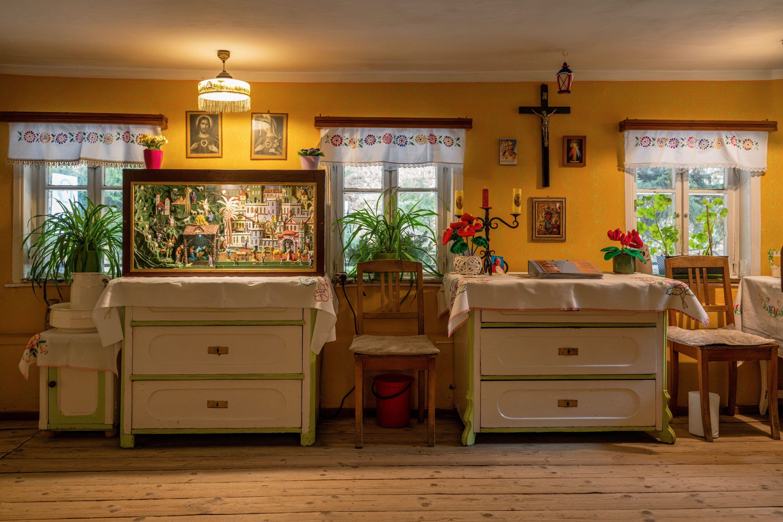 Na zdjęciu widać przytulne, żółte wnętrze z oknami, kwiatami i gablotą z ruchomą szopką ustawioną na ozdobnym stole.