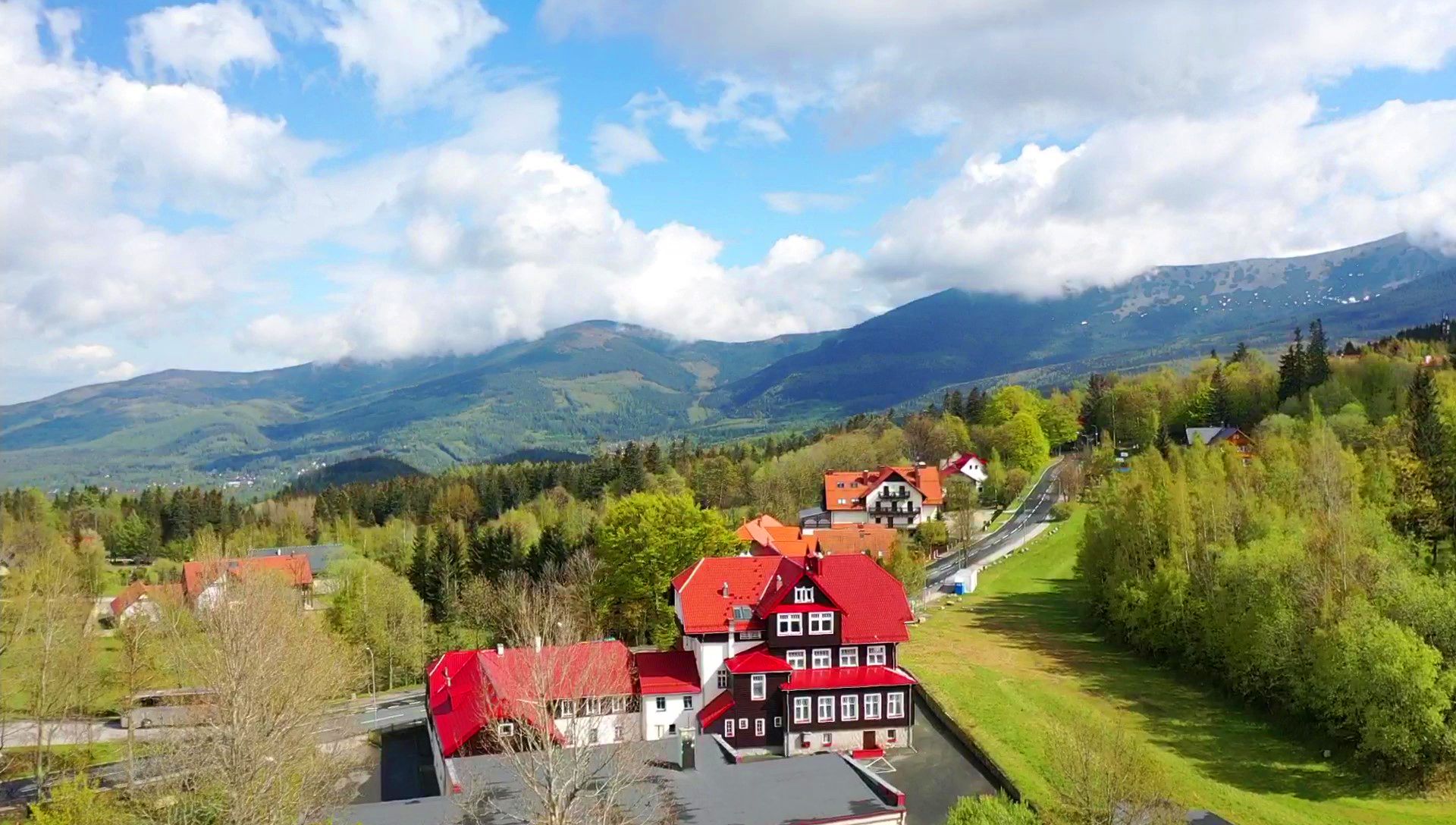 Zdjęcie z drona. Duży, charakterystyczny budynek o czerwonym dachu i ciemnej górnej kondygnacji, na tle lasu i gór. Przed wejściem znajduje się zadbane dojście z kamiennym murkiem i rabatami z kwiatami.