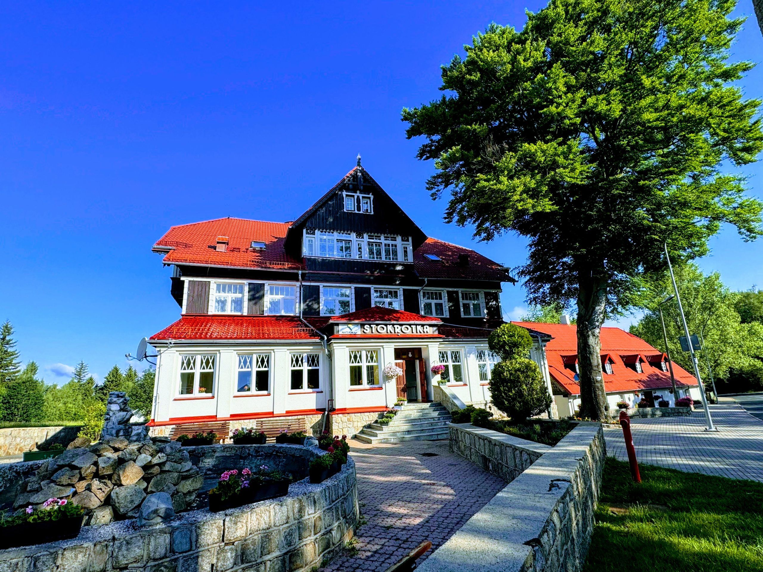 Duży, charakterystyczny budynek o czerwonym dachu i ciemnej górnej kondygnacji, otoczony zielenią i błękitnym niebem. Przed wejściem znajduje się zadbane dojście z kamiennym murkiem i rabatami z kwiatami.