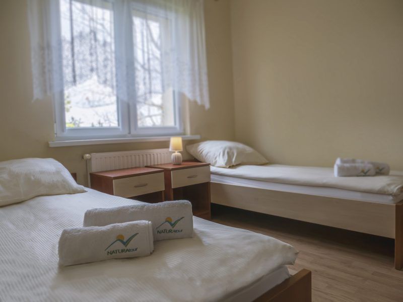 Jasny, prosty pokój z dwoma pojedynczymi łóżkami przykrytymi białą pościelą. Na łóżkach leżą ręczniki z logo „NATURA TOUR”, a całość jest oświetlona miękkim światłem lampki przy oknie.