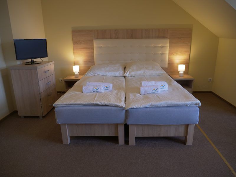 Estetyczny, prosty pokój z dużym małżeńskim łóżkiem przykrytym białą pościelą. Na łóżku leżą ręczniki z logo „NATURA TOUR”, a całość jest oświetlona miękkim światłem lampkek nocnych.