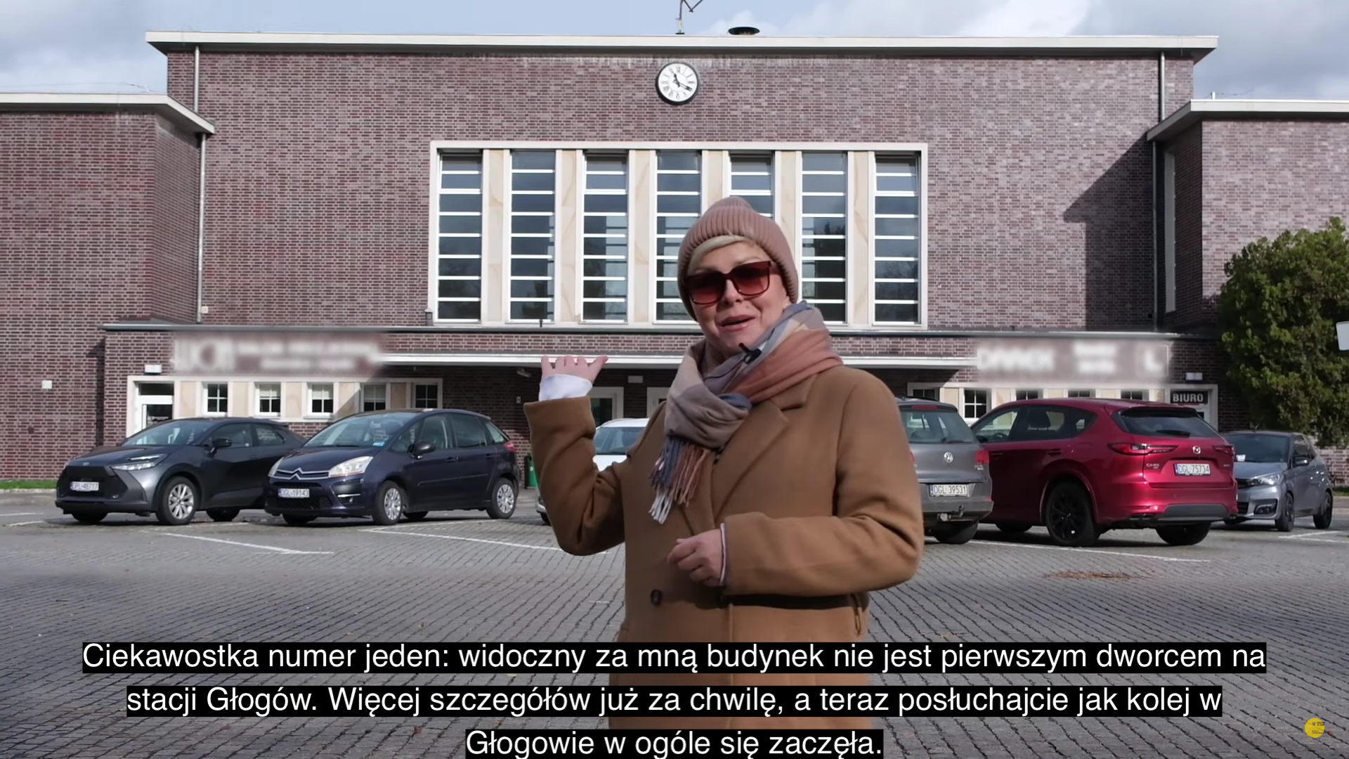 Pani Karolina Lis w płaszczu, czapce i okularach zaprasza do obejrzenia nowego odcinka na tle dworca w Głogowie