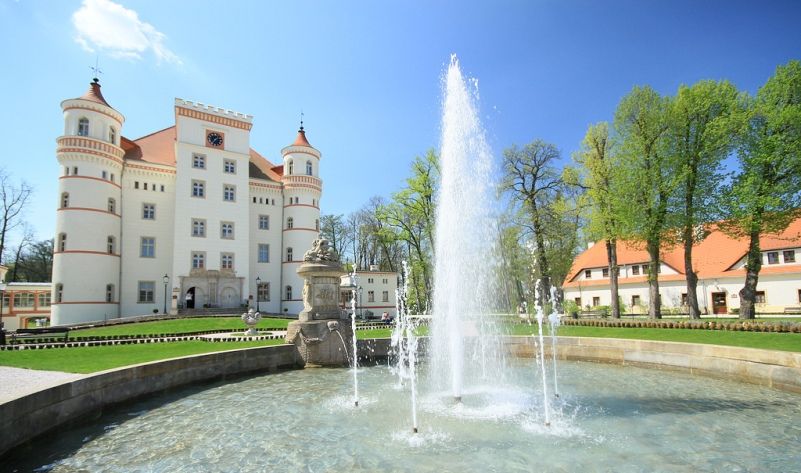 Fontanna z widocznym w tle pałacem z wieżyczkami oraz zabudowaniami folwarcznymi
