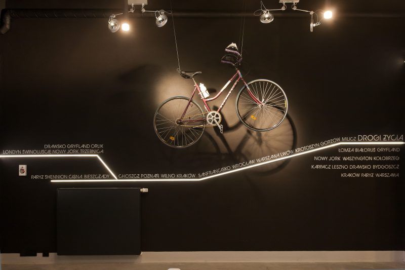Czarna ściana ekspozycyjna z podświetloną linią czasu „Drogi Życia”, na której wypisano nazwy miast. Powyżej podwieszony różowy rower turystyczny oświetlony reflektorami.