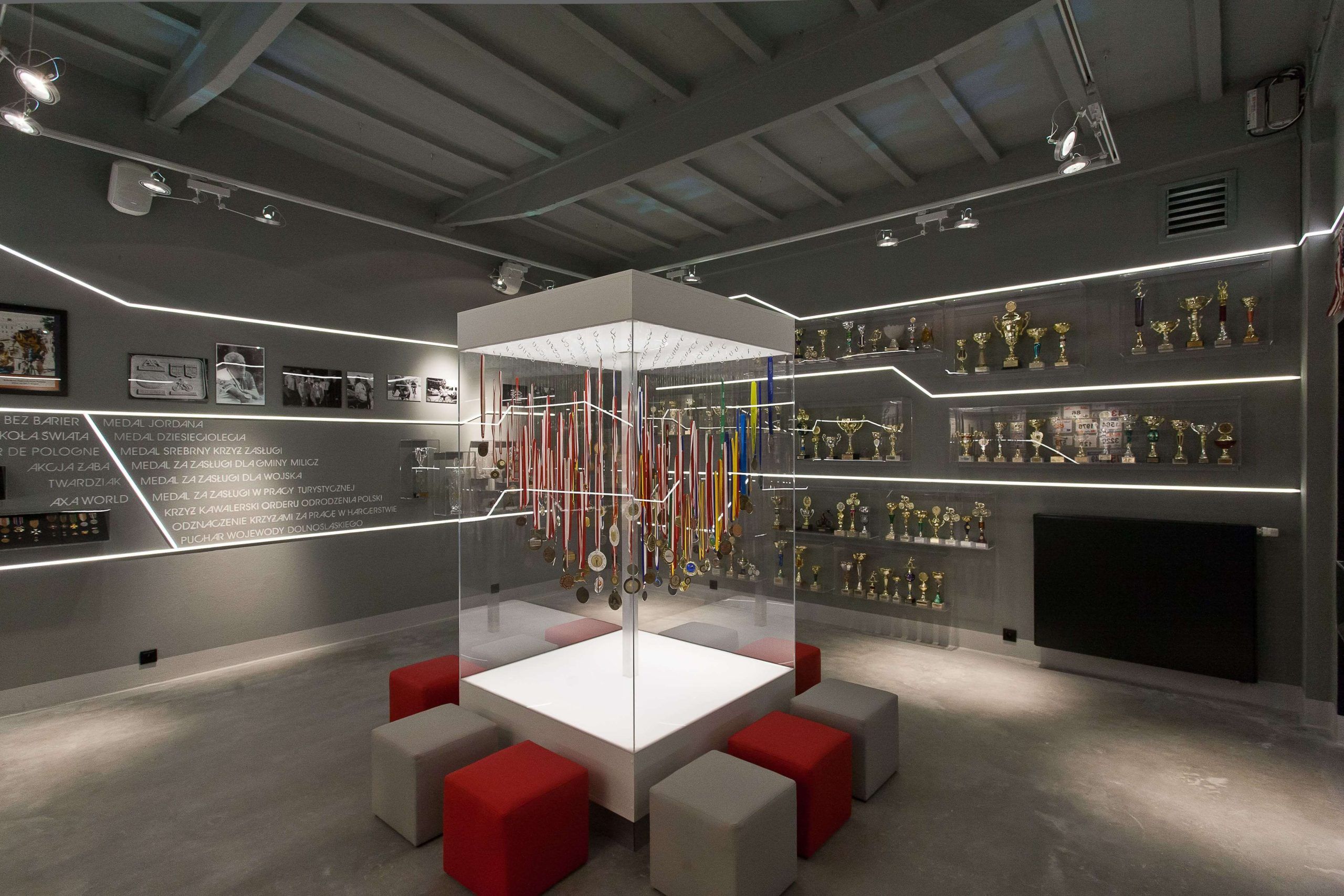 Nowoczesna sala muzealna z centralną gablotą wypełnioną medalami. Wokół gabloty znajdują się pufy do siedzenia, a na ścianach podświetlane regały z pucharami i pamiątkami.