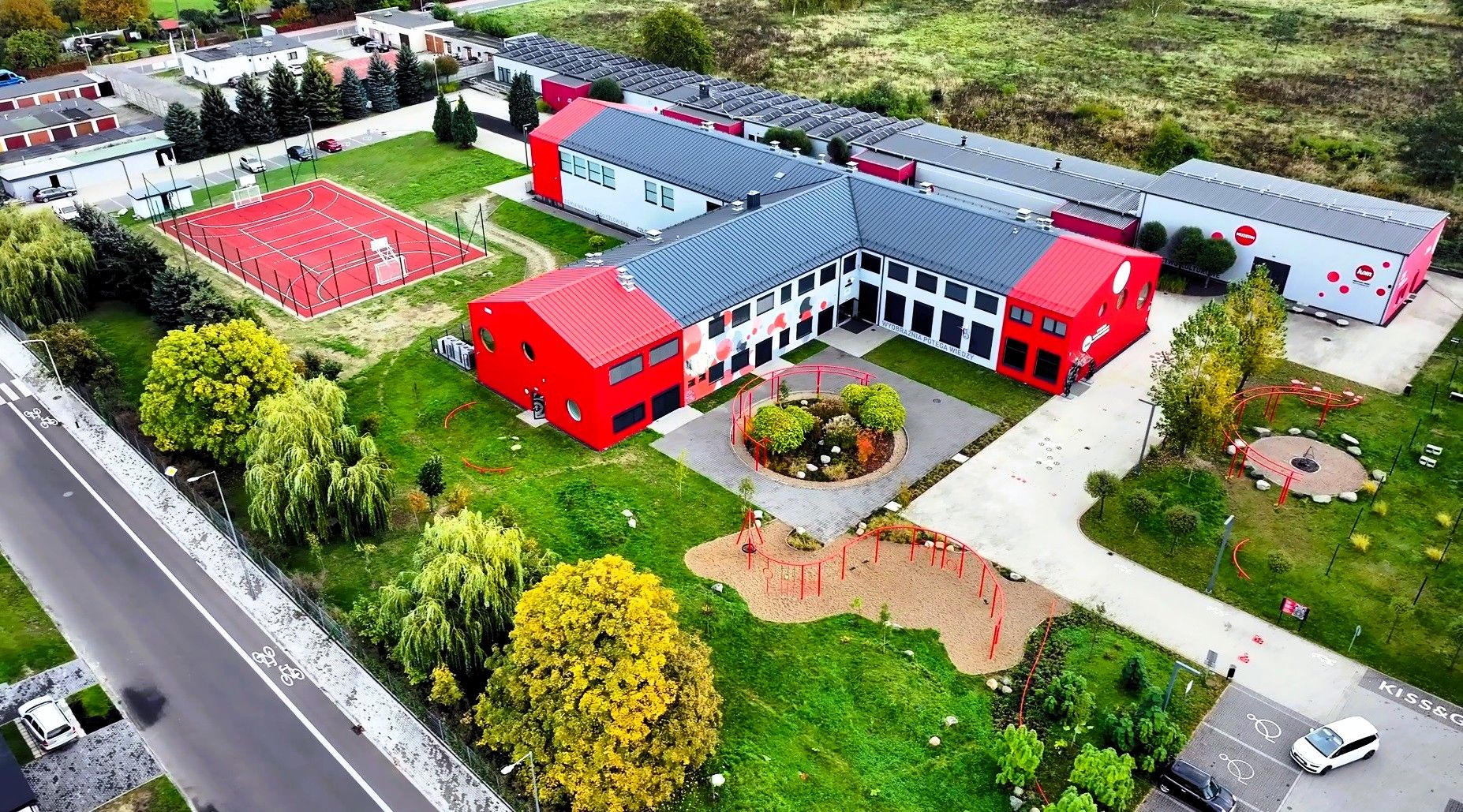 Widok z lotu ptaka na nowoczesny kompleks budynków Kreatywnego Obiektu Multifunkcyjnego w biało-czerwonej kolorystyce. Obok obiektu widoczne jest czerwone boisko wielofunkcyjne, zielony plac zabaw z kolistym klombem oraz przyległy parking i ścieżka rowerowa.