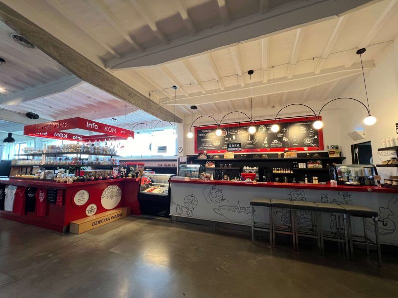 Jasne, nowoczesne wnętrze kawiarni z długim czerwonym barem i hokerami. Nad ladą wiszą lampy w kształcie łuków, a w tle widać czarną tablicę z menu oraz czerwony punkt informacyjny z napisem „info KOM shop”. Wystrój dopełniają industrialny sufit i minimalistyczne grafiki na froncie baru.