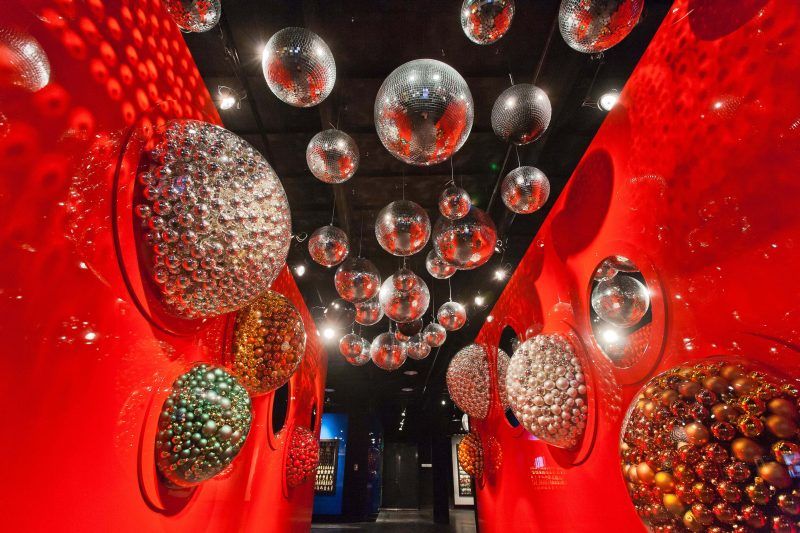 Korytarz z jaskrawoczerwonymi ścianami, w których osadzono wypukłe szklane gabloty z kolorowymi bombkami. Pod sufitem wisi kilkanaście lustrzanych kul dyskotekowych o różnych rozmiarach, które odbijają punktowe światło reflektorów.