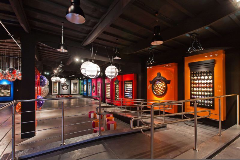 Nowoczesna sala muzealna o ciemnym industrialnym suficie, z rampą dla niepełnosprawnych i barierkami. Ściany zdobią kolorowe, podświetlane panele w kolorze pomarańczowym, czerwonym oraz niebieskim, z gablotami prezentującymi kolekcje bombek. Wnętrze oświetlają czarne wiszące lampy oraz duże, białe kule dekoracyjne.