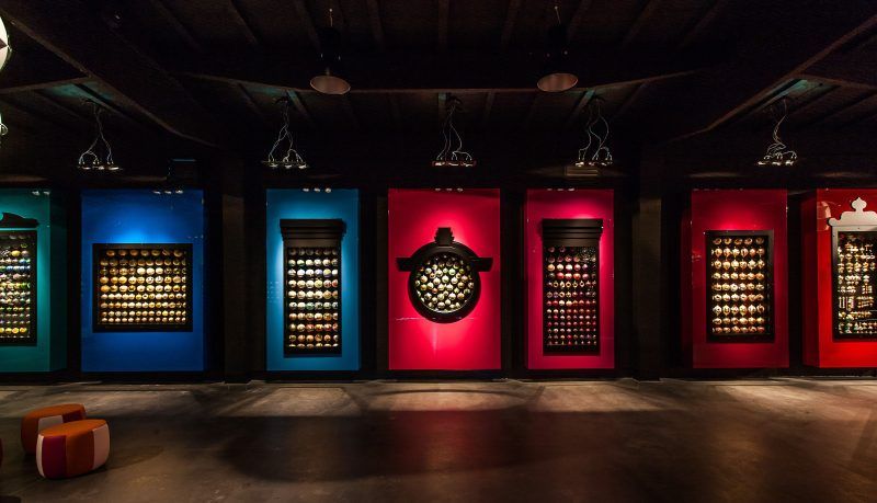 Szereg dużych, kolorowych paneli w kolorze niebieskim, różowym oraz czerownym z wbudowanymi gablotami prezentującymi kolekcje szklanych bombek. Ekspozycja znajduje się w ciemnym pomieszczeniu z punktowym oświetleniem i kolorowymi pufami na podłodze.