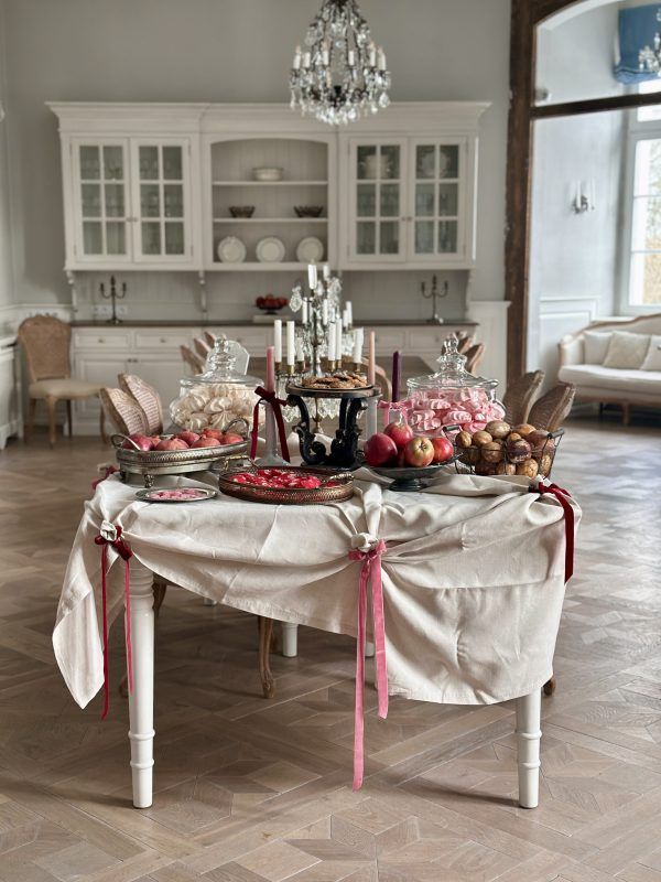 Elegancko udekorowany stół z deserami i owocami w jasnym salonie. Na białym obrusie upiętym różowymi wstążkami stoją szklane patery z bezami, misy z jabłkami i orzechami oraz wysokie świece. W tle widoczny klasyczny biały kredens i kryształowy żyrandol.