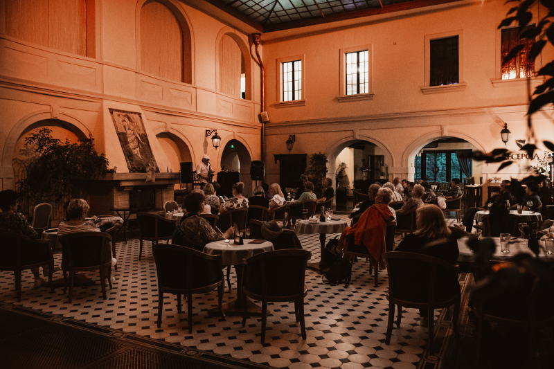 Przestronny hotelowy hol z wysokim sufitem i arkadami podczas wydarzenia kulturalnego. W centralnej części ustawiono stoliki z krzesłami, przy których siedzą ludzie, najpewniej wysłuchując koncertu, bądź wykłądu. Po bokach widać recepcję i kominek.