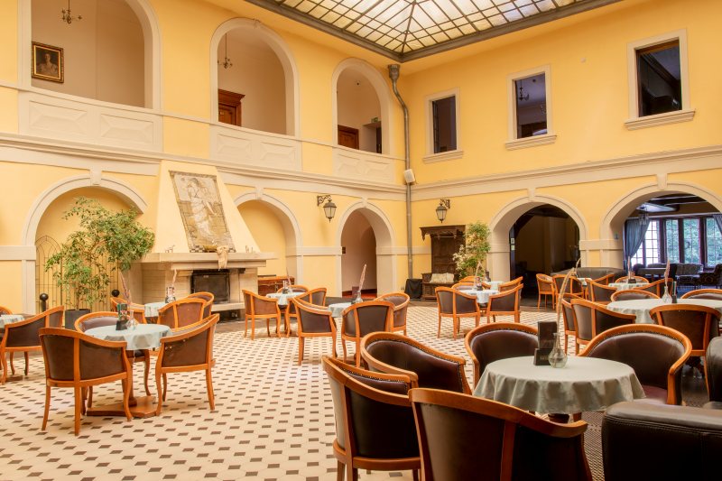 Przestronny hotelowy hol z wysokim sufitem i arkadami tworzy elegancką, klasyczną przestrzeń do wypoczynku. W centralnej części ustawiono stoliki z krzesłami, a po bokach widać recepcję, kominek oraz miejsca do siedzenia.