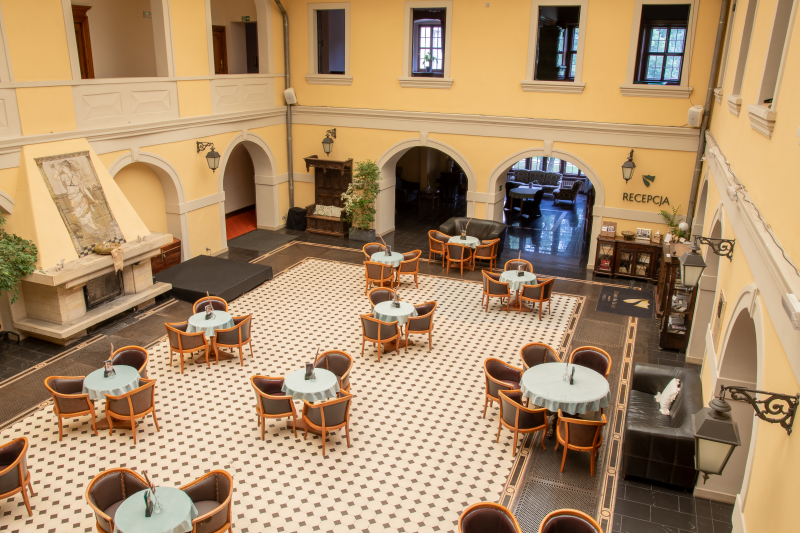 Przestronny hotelowy hol z wysokim sufitem i arkadami tworzy elegancką, klasyczną przestrzeń do wypoczynku. W centralnej części ustawiono stoliki z krzesłami, a po bokach widać recepcję, kominek oraz miejsca do siedzenia.