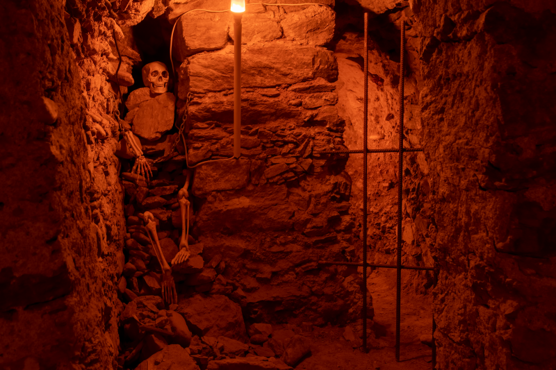 Zdjęcie przedstawia kamienne podziemia o surowych ścianach, oświetlone ciepłym, czerwonym światłem. W jednym z narożników znajduje się szkielet przymocowany łańcuchami do ściany, co nadaje miejscu mroczny, tajemniczy charakter.