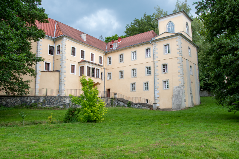 Na zdjęciu widoczny jest jasny, historyczny budynek o czerwonym dachu, otoczony zielenią i rozległym trawnikiem. Obiekt znajduje się w spokojnym, parkowym otoczeniu, a nad nim widać chmury.