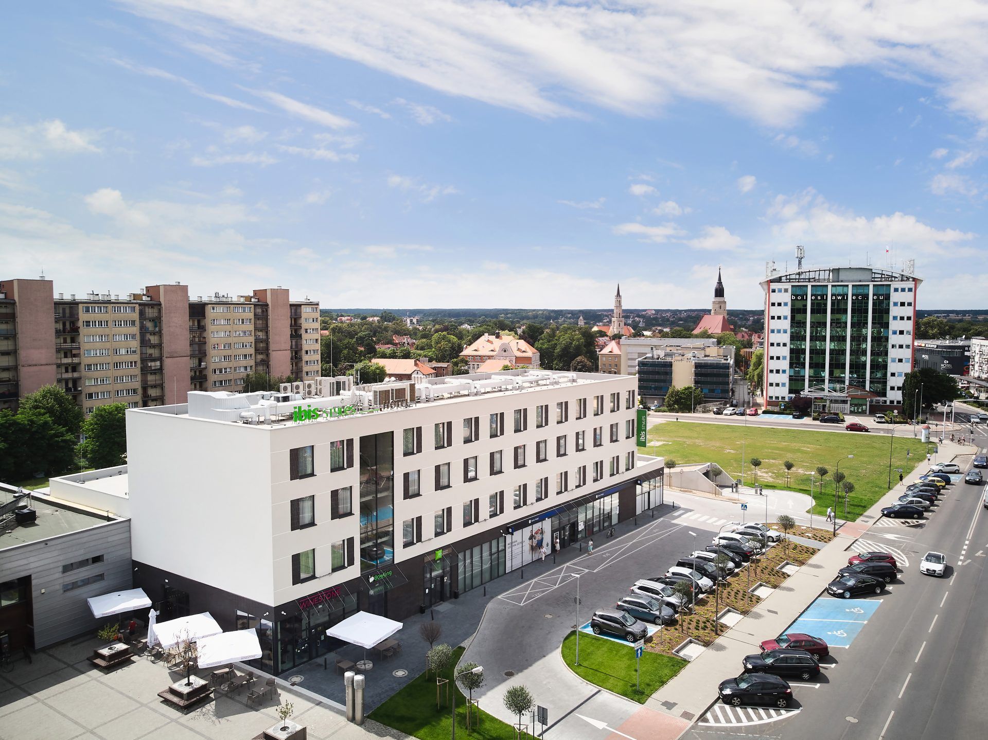 Nowoczesne budynki, w tym hotel z charakterystycznym zielonym szyldem oraz fragment parkingu z samochodami.