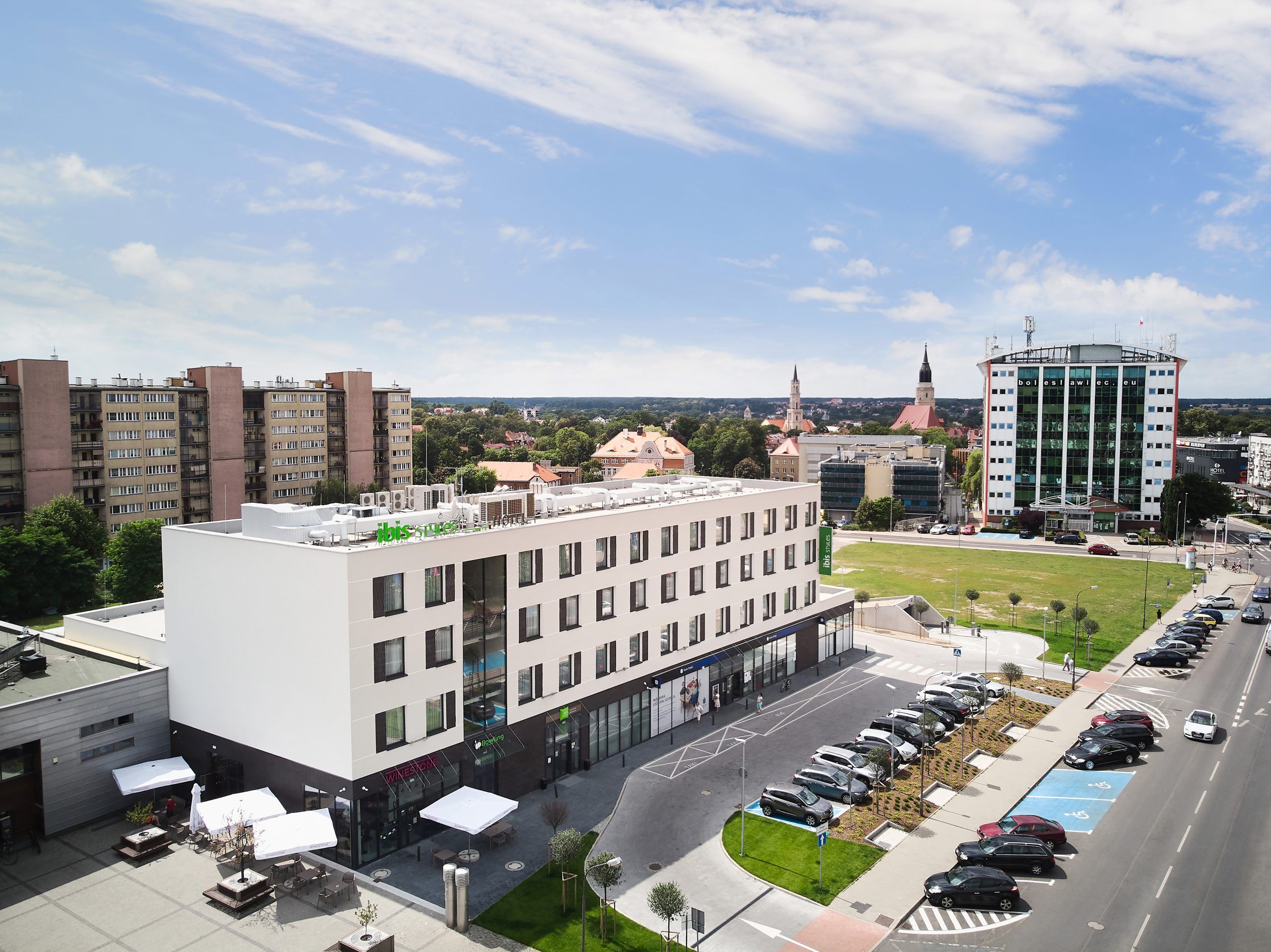 Nowoczesne budynki, w tym hotel z charakterystycznym zielonym szyldem oraz fragment parkingu z samochodami.