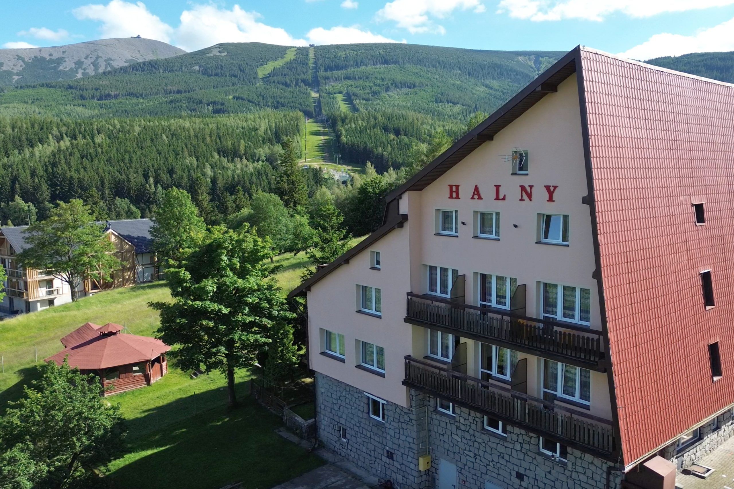 Na zdjęciu widoczny jest budynek ośrodka wypoczynkowego „Halny”, położony w górskim otoczeniu u podnóża Karkonosze. W tle widać zalesione stoki oraz charakterystyczną trasę prowadzącą na szczyt Śnieżka, co podkreśla malowniczy, górski charakter miejsca.