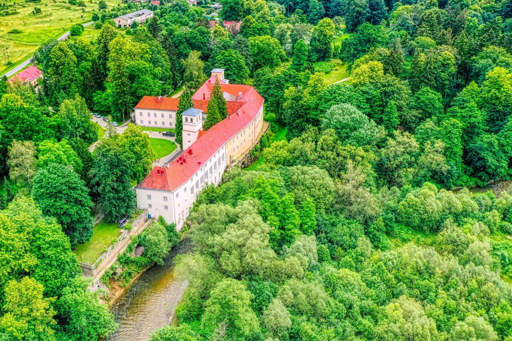 Na zdjęciu z lotu ptaka widać rozległy budynek z czerwonym dachem, położony wśród gęstej zieleni drzew. Obiekt stoi tuż nad zakolem rzeki,