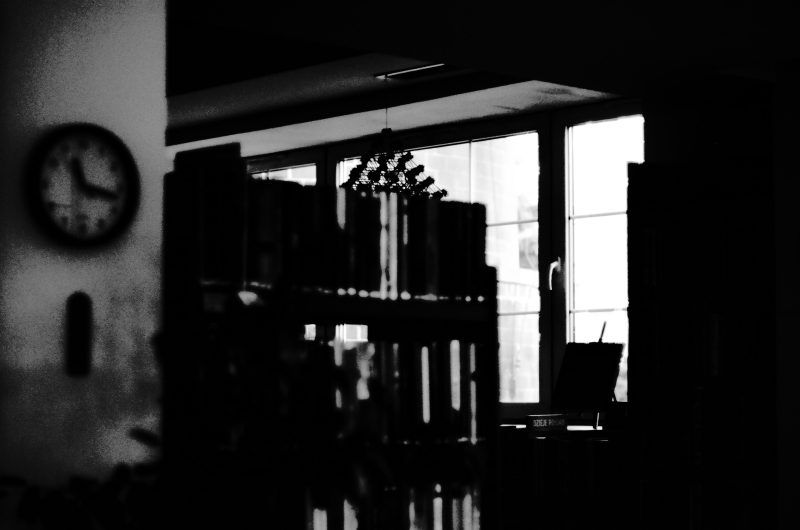 Czarno-biała, kontrastowa fotografia przedstawia mroczne wnętrze biblioteki lub czytelni, gdzie głębokie cienie zderzają się z jasnym światłem wpadającym przez okno. Na pierwszym planie, po lewej stronie, widoczny jest ziarnisty zarys ściany z zawieszonym wysoko okrągłym zegarem. Centralną część zajmuje pogrążony w mroku regał, którego zawartość tworzy rytm pionowych, ciemnych linii. W tle, na tle jasnych szyb wielodzielnego okna, rysują się ostre sylwetki wiszącej lampy oraz stojaków na książki umieszczonych na blacie. Całość ma surowy, ziarnisty i bardzo nastrojowy charakter, przypominający kadr ze starego filmu.