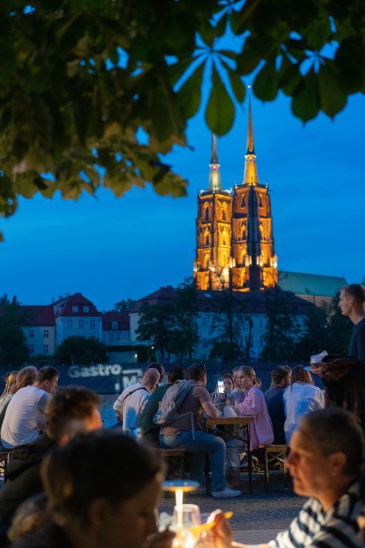 widok pionowy na wrocławską katedrę i ludzi siędząccych przy stolikach po zachodzie słońca, podczas wydarzenia gastro miasto