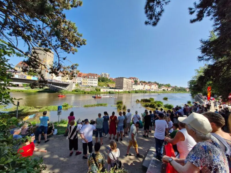 Una folla di spettatori si è radunata attorno allo stagno durante un evento all'aperto nelle Bory Dolnośląskie, osservando gli edifici dall'altra parte dell'acqua.