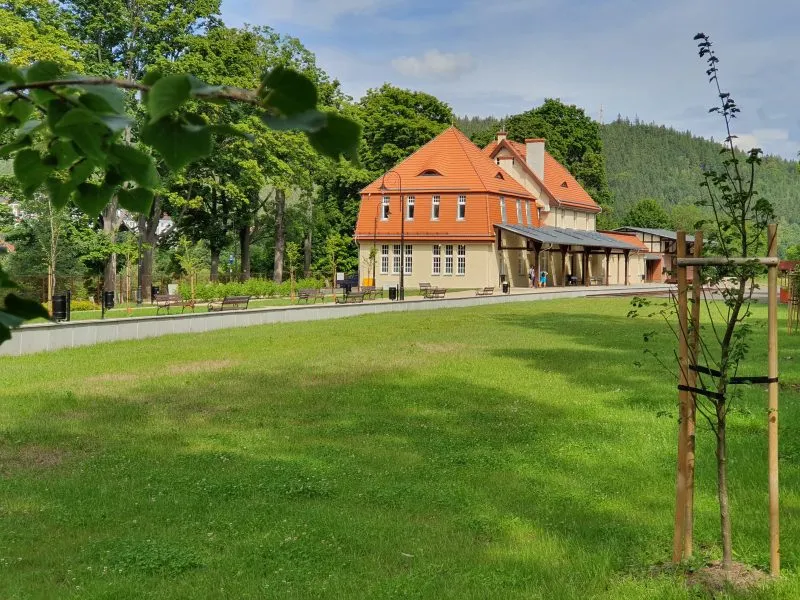 L'edificio rinnovato della Stazione della Cultura, con il tetto arancione e la facciata chiara, sorge accanto a un ampio prato, circondato da giovani alberi e colline boscose.