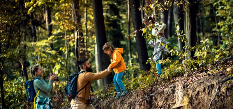 I genitori aiutano i bambini a salire su un ripido pendio boscoso, porgendo loro le mani e percorrendo insieme il sentiero tra alberi e rocce.