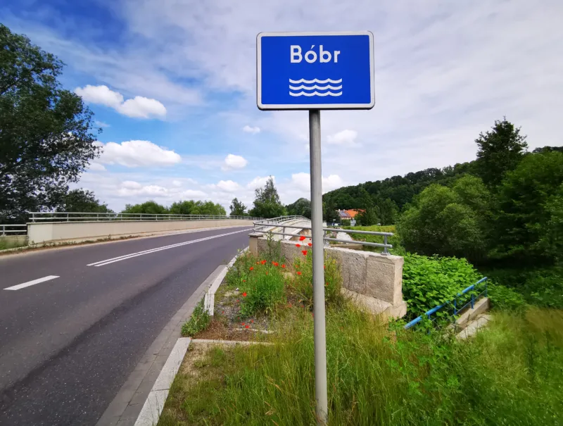 Zdjęcie przedstawia niebieski znak drogowy z napisem Bóbr stojący przy asfaltowej drodze