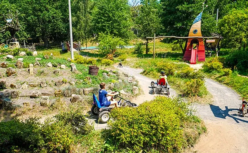 I bambini guidano piccoli veicoli fuoristrada lungo i sentieri di un verde campeggio, passando accanto a giardini rocciosi, cespugli e una casetta di legno.