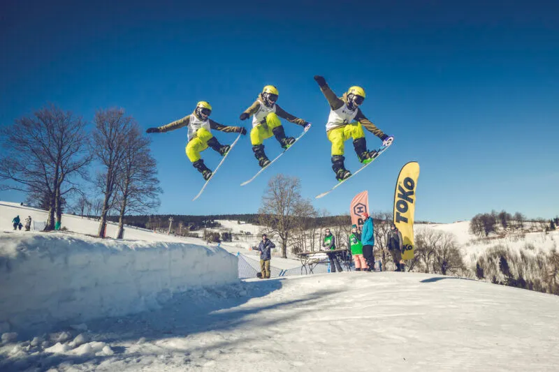 Uno snowboarder esegue un salto elevato sopra una pista innevata nella Terra di Kłodzko, mostrato in tre fasi successive dell'evoluzione sullo sfondo di un cielo azzurro.