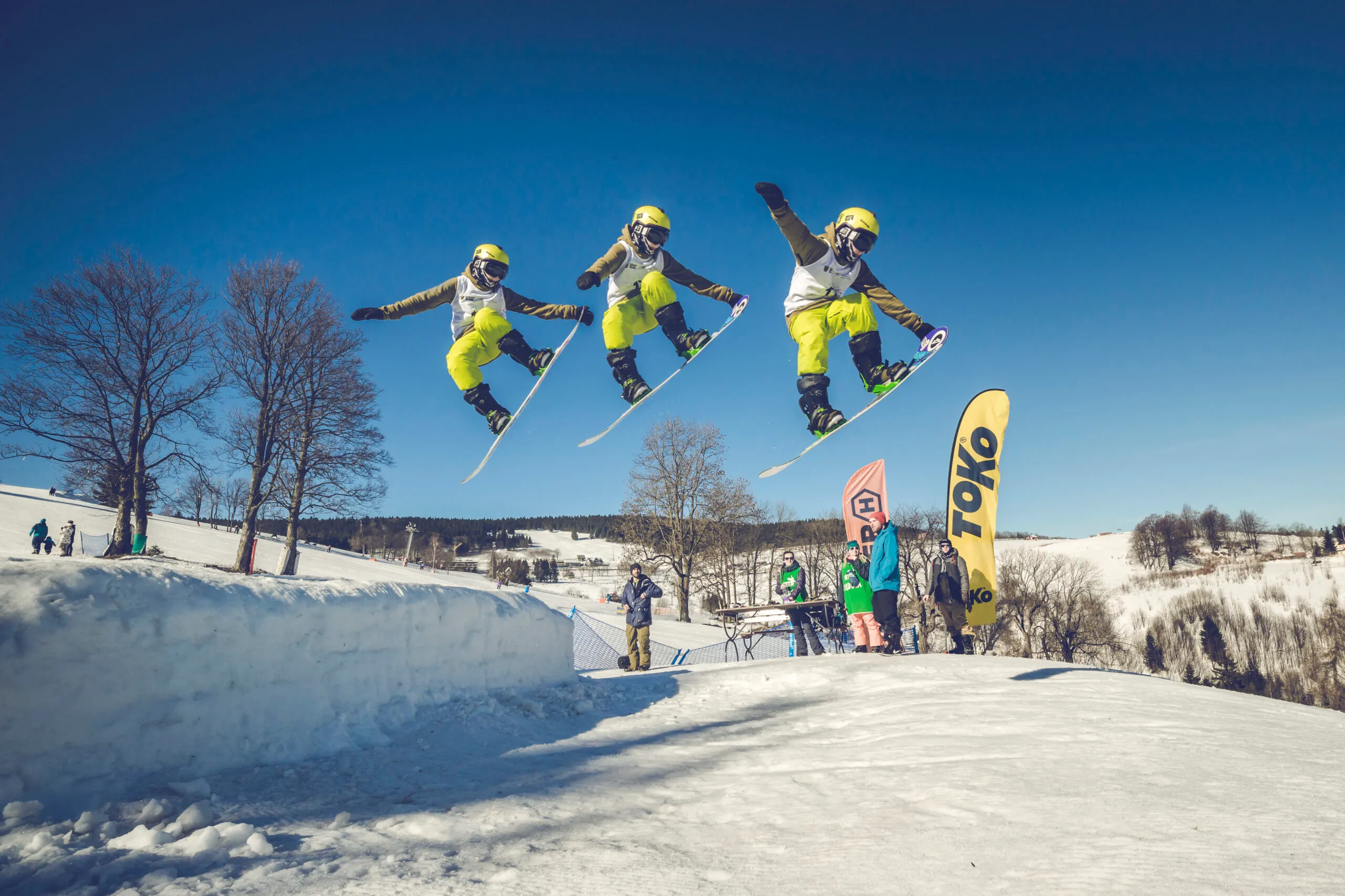 A snowboarder performs a high jump over a snowy slope in the Kłodzko Valley, shown in three successive phases of the evolution against a blue sky.